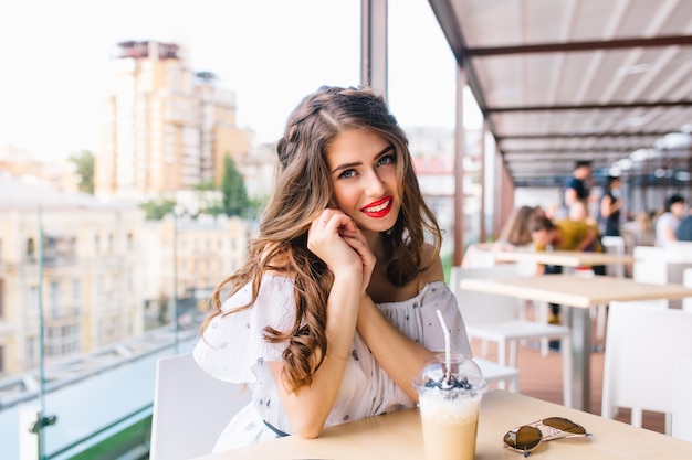 Горизонтальный портрет красивой девушки с длинными волосами, сидя за столом на террасе в кафе. Она носит белое платье с открытыми плечами и красной помадой. Она дружелюбно улыбается в камеру.