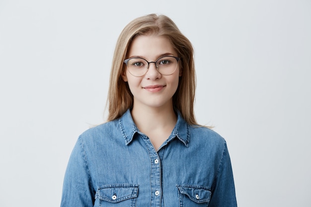 행복 한 젊은 쾌적 한 찾고 여성 미소의 가로 세로 스트레이트 금발 머리와 데님 셔츠와 세련 된 안경을 착용, 긍정적 인 표현 포즈