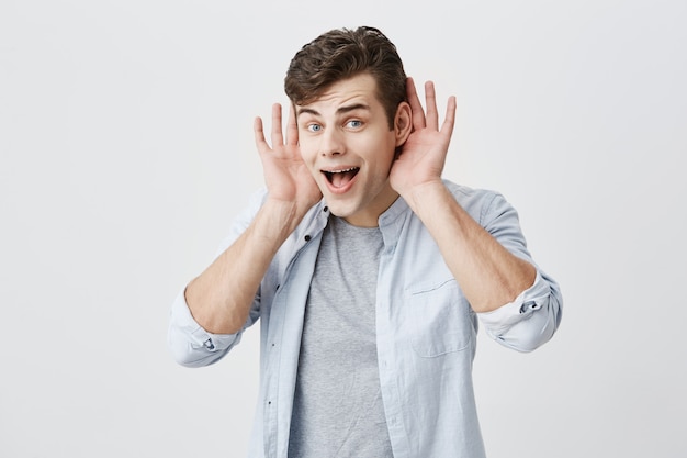 Horizontal portrait of surprised caucasian young man being astonished with big sales prices, has shocked look, looks with eyes popped out and jaw dropped, holding palms open behind his ears.