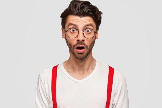 Горизонтальный портрет удивленного небритого парня с отвисшей челюстью, не может поверить в свою неудачу, носит круглые очки, изолирован на белой стене. Удивленный кавказский мужчина на что-то реагирует