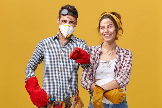 Horizontal portrait of young foreman wearing safety eyewear, mask and red gloves holding tool belt pointing with finger standing near her female colleague having smiles on their faces