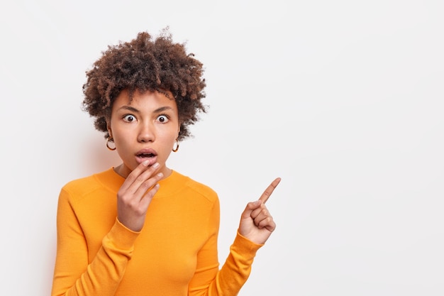 Horizontal shocked speechless woman with Afro hair keeps jaw dropped indicates away on blank space says check out something unusual wears long sleeved orange jumper isolated over white wall