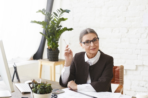 Horizontal shot of 50 year old woman architect in formal wear holding pencil while doing paperwork in light office, checking technical drawings, having serious expression. Architecture and engineering