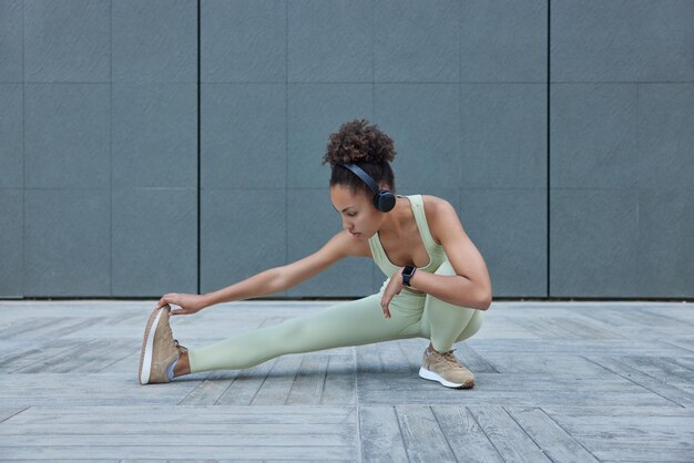 Horizontal shot of active slim woman stretches legs demonstrates her flexibility listens audio track in wireless headphones wears tracksuit