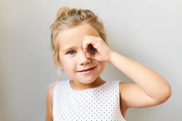 Horizontal shot of adorable pretty girl in beautiful dress posing  looking through binocular made of her hand, connecting thumb with fore finger. Cute funny female child having fun, spying