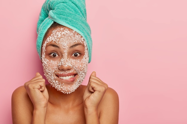 Horizontal shot of adorable young woman bites lips, clenches fists with satisfaction, looks gladfully, has face covered with sea salt mask cares about complexion stands bare shoulders indoor