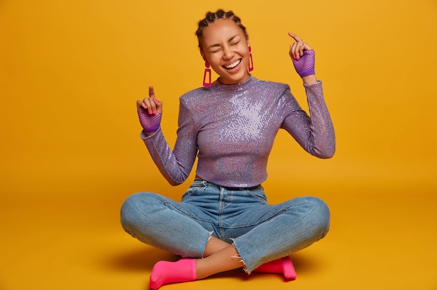 Horizontal shot of amused carefree dark skinned girl sits crossed legs and laughs, raises arms, has group meeting, hears something very funny, closes eyes from positiveness, isolated on yellow wall