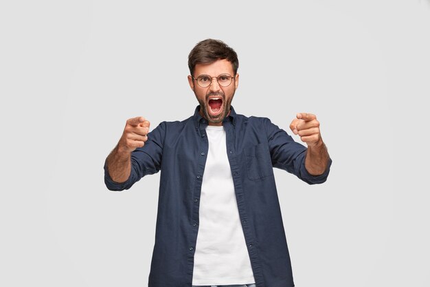 Горизонтальный снимок разгневанного разъяренного человека с раздраженным выражением лица, который кричит на кого-то, указывает указательными пальцами на камеру