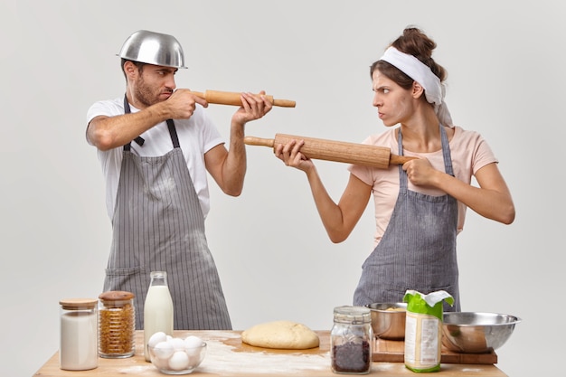 Horizontal shot of angry husband and wife feel like opponents, shoot at each other with rolling pins, cook together at home, make dough with flour, prepare delicious pastry, do bakery. Kitchen fight