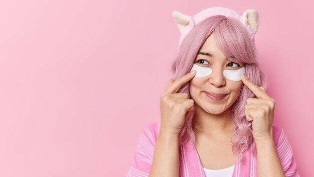 Foto gratuita l'immagine orizzontale di una donna asiatica con punti di capelli tinti di rosa sulle macchie di bellezza sotto gli occhi per rimuovere le rughe guarda pensierosamente via indossa una fascia morbida isolata su uno spazio vuoto di sfondo rosa