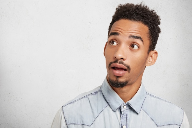 Horizontal shot of astonished hipster male with trendy hairstyle, hazel eyes, beard and mustache looks surprisely aside,