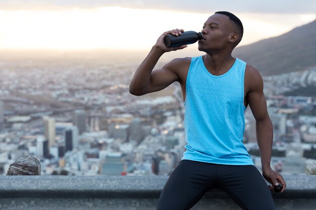 Horizontal shot of attractive active young male with muscular hands, holds bottle of water, rests after intensive jog, wears sportsclothes, sits on road sign