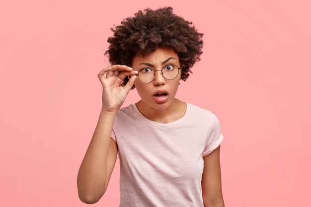 Horizontal shot of attractive African American female with scrupulous expression, looks with indignation through eyewear, opens mouth, tries to read something from distance, poses indoor alone