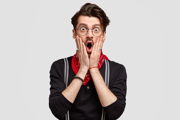 Horizontal shot of attractive bearded young man covers cheeks with palms, stares with shocked expression