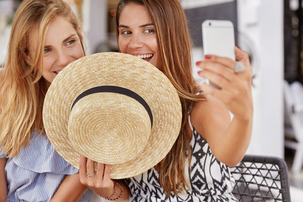 Horizontal shot of attractive beautiful women hide behind straw hat, make selfie with phone, share their photos in social networks online. Delighted lesbian couple make photo of themselves