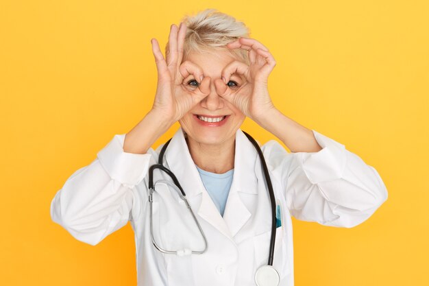 Horizontal shot of attractive cheerful middle aged female medical worker wearing white coat and stethoscope around neck having fun, showing positive attitude, making binoculars with fingers, smiling