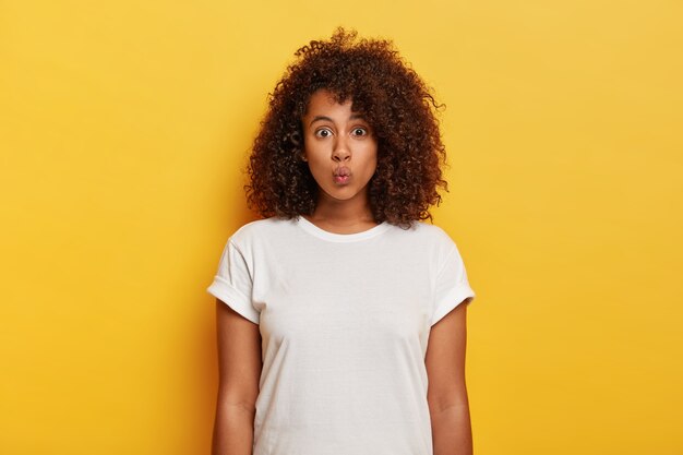 Horizontal shot of attractive flirty woman makes lips folded, waits for passionate kiss from boyfriend, has lovely outlook, curly hairstyle, wears white t shirt, models against yellow wall.