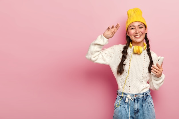 魅力的な混血の女性の水平方向のショットは、現代の携帯電話を保持し、踊り、首にヘッドフォンを着用し、ピンクの背景、コピースペースに対してポーズをとる