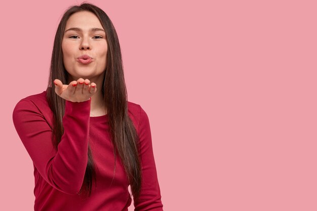 Horizontal shot of attractive woman blows air kiss, keeps palm near folded lips, has long hair