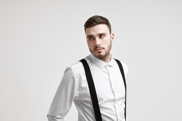 Horizontal shot of attractive young bearded Caucasian employee dressed in elegant suit posing in studio, looking aside with serious pensive expression, thinking over business ideas and plans