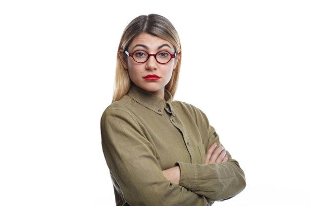 Horizontal shot of attractive young female with blonde loose hair and red lips keeping arms folded, her look and posture expressing dislike or disagreement about some business decision or idea