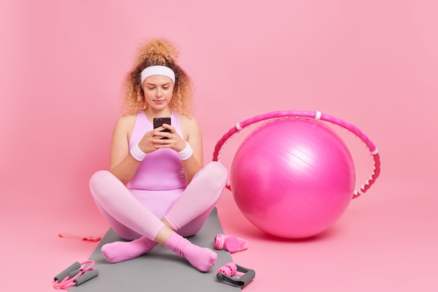 Horizontal shot of beautiful curly female model in good shape sits crossed legs on fitness mat uses mobile phone checks burned calories in special app uses sport equipment 