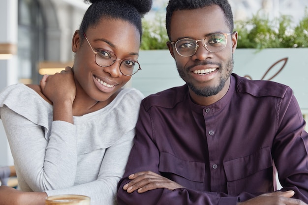 Horizontal shot of beautiful dark skinned female with cheerful expression, happy to meet her best African American friend, sit in outdoor cafe