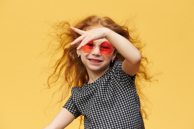 Free photo horizontal shot of beautiful happy trendy little girl with curly ginger hair enjoying dancing,  with cheerful broad smile, wearing sunglasses. music, dance, fun and children concept