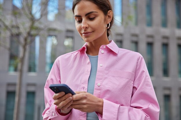 Horizontal shot of beautiful millennial girl uses mobile phone in city for finding route explores new places of interest wears pink shirt poses on blurred building 
