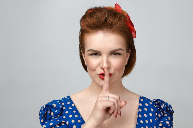 Horizontal shot of beautiful pin up girl in vintage clothes saying shush, holding index finger at her lips and smiling mysteriously, asking for silence, making suprise party for her husband