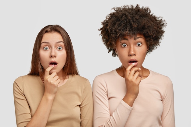 Free photo horizontal shot of beautiful scared two multiethnic women with stunned expressions, keep hands near widely opened mouthes, react on fake news, stand next to each other, isolated over white wall.