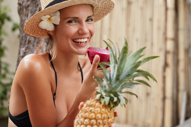 Horizontal shot of beautiful smiling woman with broad shining smile, wears summer hat and bathing suit, holds tropical fruits, enjoys unforgettable summer rest, spends leisure time in tropics