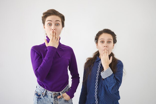 Horizontal shot of beautiful stylish 20 year old woman and her teenage sister expressing astonishment and full disbelief, covering mouths