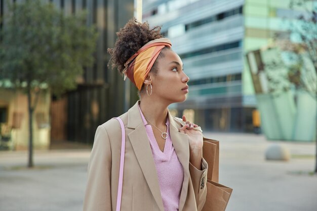 Horizontal shot of beautiful woman focused away with thoughtful expression carries paper shopping bags poses in downtown against modern buildings.