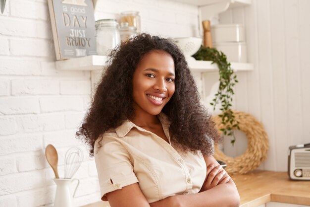 Horizontal shot of beautiful young black mulatto woman in beige shirt crossing arms on chest, looking  with confident toothy smile, posing indoors against stylish kitchen interior 