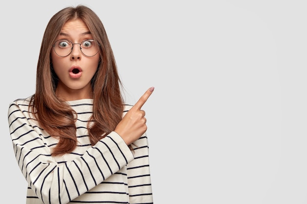 Horizontal shot of beautiful young Caucasian female with surprised expression, points at upper right corner, dressed in striped sweater, wears round spectacles, isolated over white wall