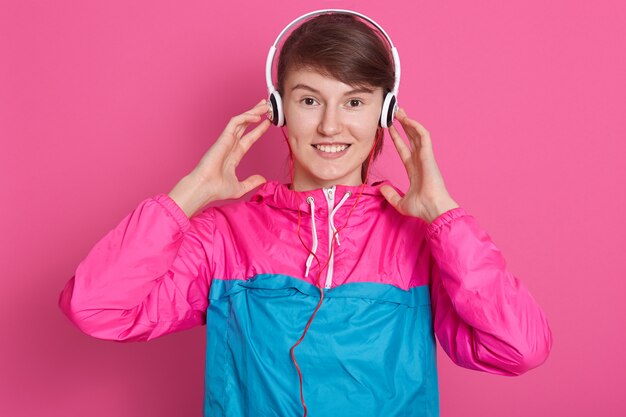 スポーツウェアの美しい若い白人の女の子の水平方向のショットは、ヘッドフォンで音楽を聴いて楽しむ、ピンクの壁を越えて耳に手を当てています。フィットネス、スポーツ、健康的なライフスタイルのコンセプト