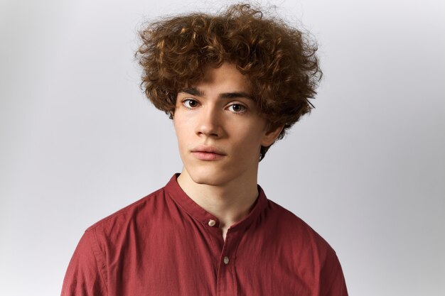 Horizontal shot of brown eyed cute young Caucasian male with wavy hair looking at camera with curious facial expression listening to you attentively.