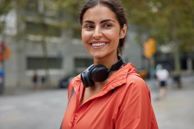 Horizontal shot of brunette woman has cheerful facial expression leads active lifestyle dressed in windbreaker wears stereo headphones poses outside against blurred background Sport concept