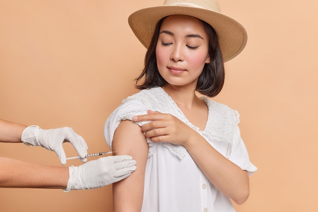 ブルネットの若いアジアの女性の水平方向のショットは、フェドーラ帽と白いドレスを着て肩に接種を取得します