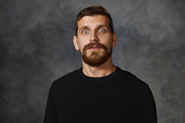 Horizontal shot of bug eyed handsome adult male with stubble posing wearing black sweatshirt widening his eyes, having shocked panic look, showing full disbelief, terror or fright