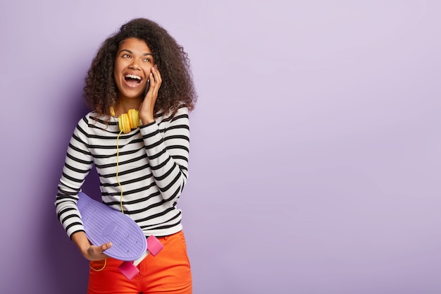 Horizontal shot of carefree optimistic lady with curly dark hair, calls best friend, has happy mood