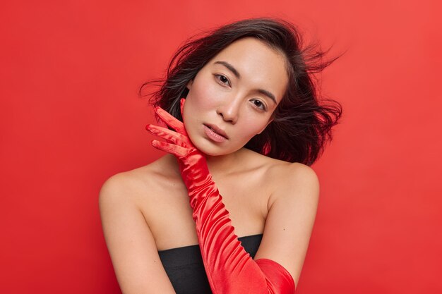 Horizontal shot of charming Asian woman with natural beauty touches face gently has dark hair floating in air wears black dress long gloves isolated over vivid red wall