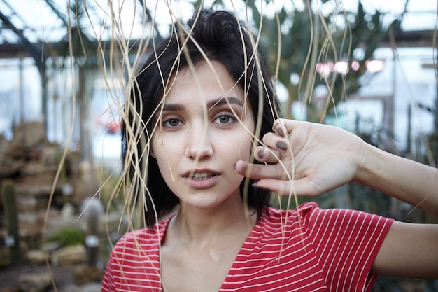 Free photo horizontal shot of charming bob-haired young female shopping in garden center, standing against blurred background of plants with straw stalks hanging down in front of her. gardening and agriculture