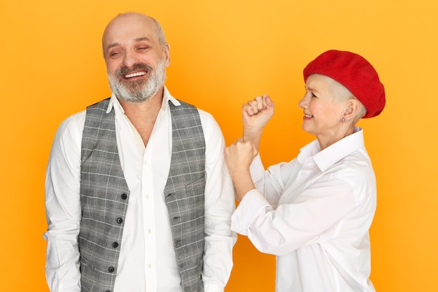 Horizontal shot of cheerful bearded bald senior man in elegant clothes smiling having careless facial expression, his anrgy furious middle aged woman being mad, punching him in shoulder, squabbling