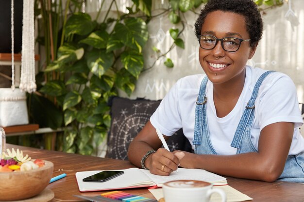 Horizontal shot of cheerful black woman wears spectacles writes list to do in notebook or personal diary