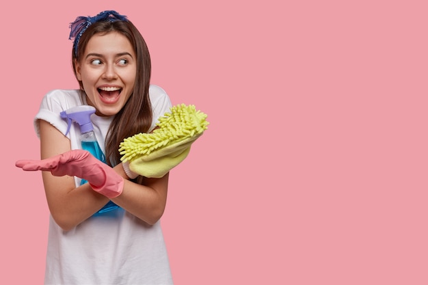 Horizontal shot of cheerful brunette woman crosses hands, carries sponge and detergent