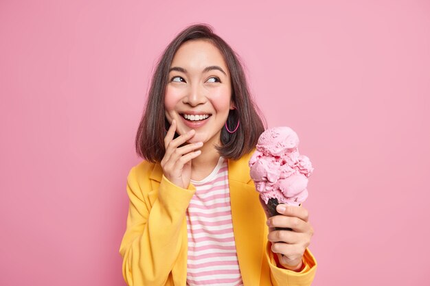 Horizontal shot of cheerful brunette young Asian woman looks away gladfully holds delicious cold ice cream in waffle wears elegant clothes enjoys summer holidays isolated over pink wall.