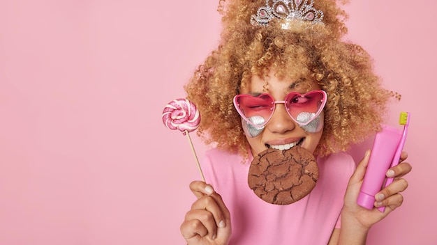 Free photo horizontal shot of cheerful curly haired woman eats tasty cookie winks and looks gladfully aside holds lollipop in one hand and teeth care product in other isolated over pink background empty space