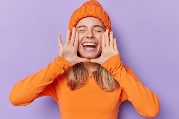 Horizontal shot of cheerful millennial girl keeps hands near opened mouth whispers good news keeps eyes closed wears knitted orange hat and poloneck feels very glad isolated over purple background